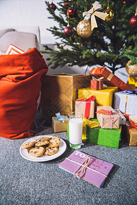在礼物和圣诞树附近的地板上放着一杯牛奶饼干和给图片