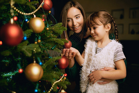 美丽的女孩和她的母亲在圣诞树的fir树上看着一个装饰图片
