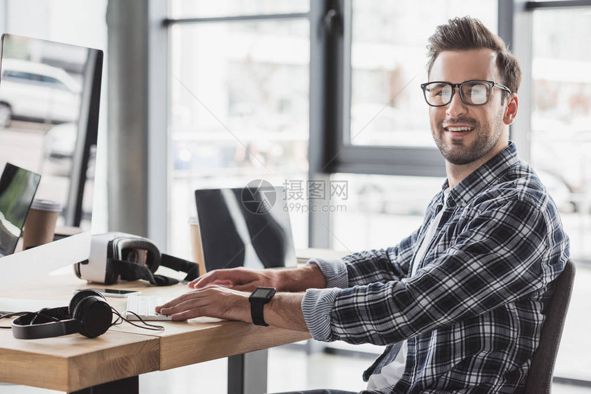 身戴眼镜的英俊青年男子在与台式电脑和笔记本电脑一起工作时图片