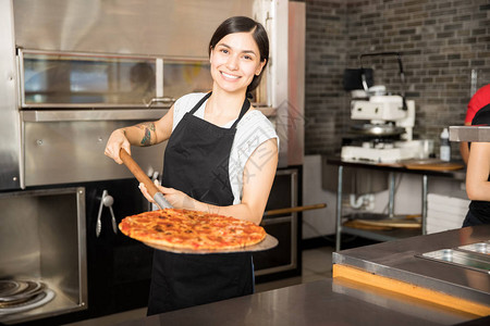 在比萨饼店工作的快乐的年轻女士展示新鲜烤面包披萨站在图片
