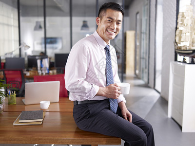 一个亚洲商人拿着咖啡杯坐在办公室的桌子上笑图片