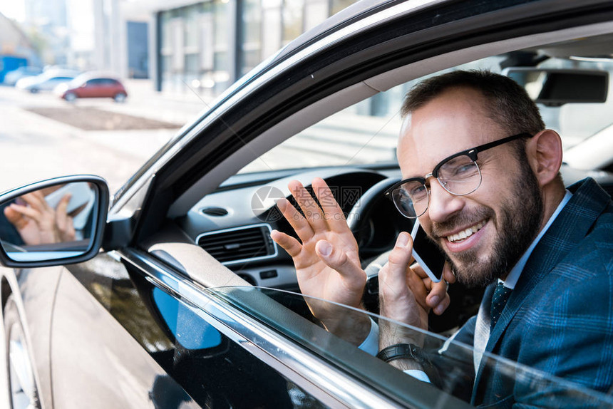 在汽车中挥手时用智能手机说话的眼镜上的快乐的胡须商人有图片