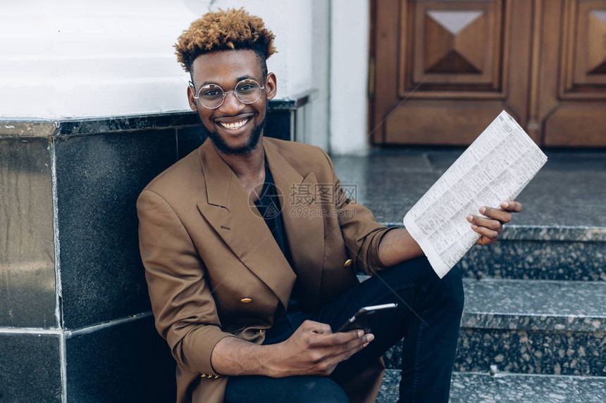 一名身穿夹衣有智能手机和关于办公大楼背景的报纸的非裔美国人图片