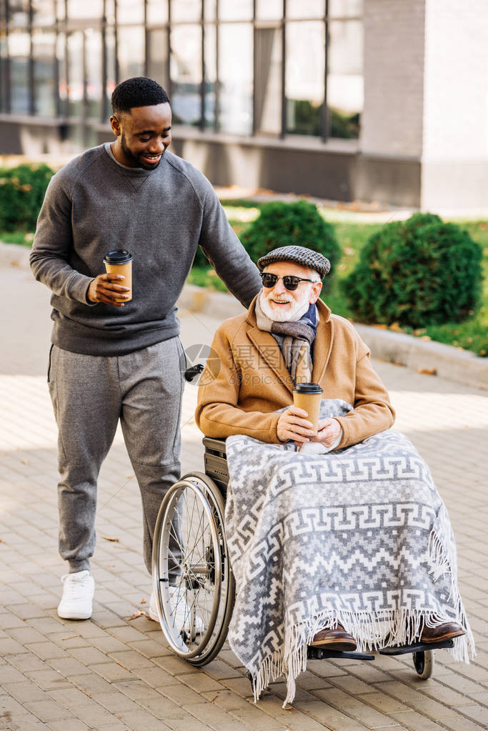 坐在轮椅上笑着微笑的老年残疾人与格子和非洲裔美国人男子一起在街上骑车图片