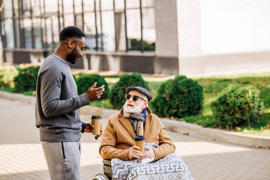坐在轮椅上笑着微笑的老年残疾男子与格子和非洲裔美国人男子一起在街上用纸杯图片
