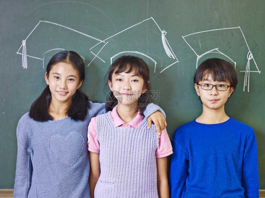 三名亚裔小学生站在粉笔拖着的博士帽下面的黑板前图片