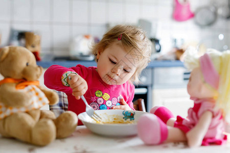 可爱的女婴吃叉子蔬菜和意大利面小孩喂养和玩具娃和毛绒熊可爱的蹒跚学步图片