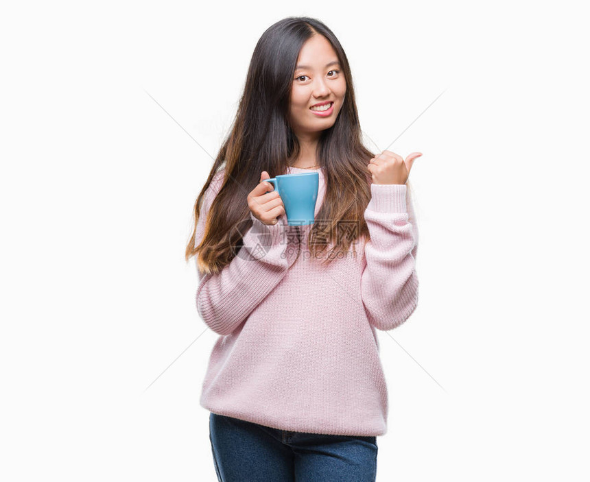 年青的亚洲女人在孤立的背景点上喝咖啡然后用拇指举起手笑着图片
