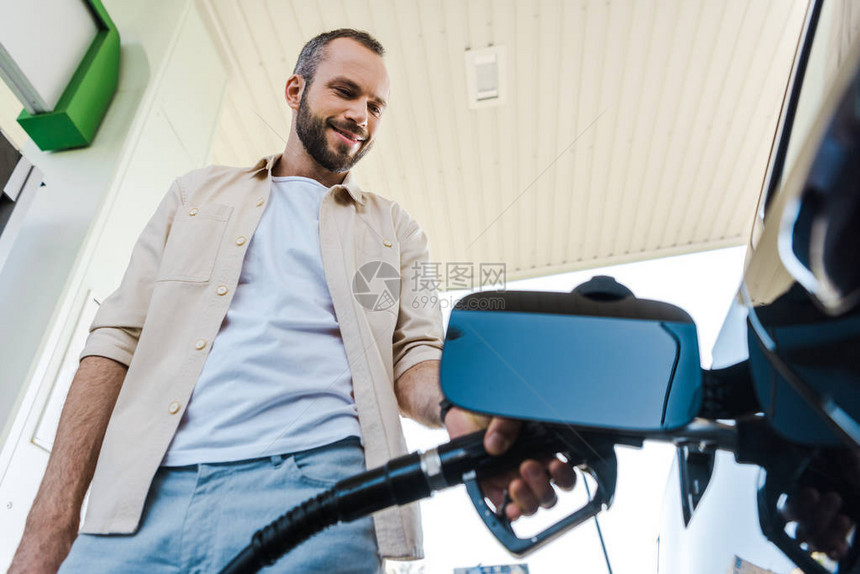 在加油站为汽车加油时持有燃料泵的快乐男子低角度视图图片