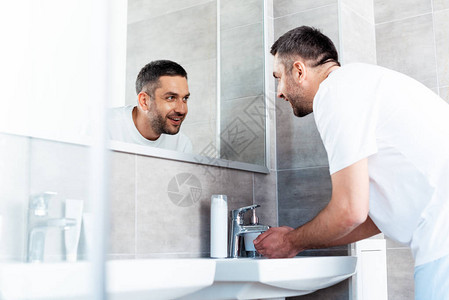 长着英俊的笑脸男人在照镜子和洗手在早晨例行活动期间图片