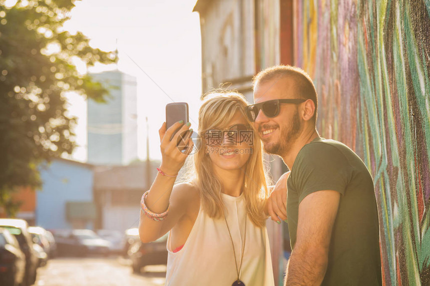 一对快乐的情侣带着智能手机自拍站在街图片