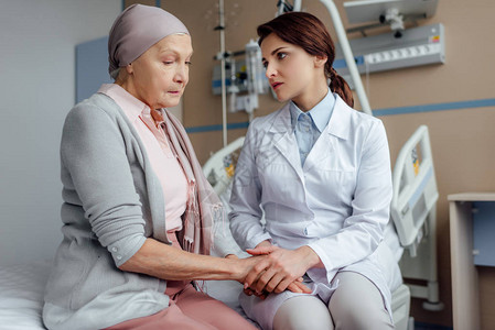 与在医院患癌症的手帕上戴头巾的高级妇女握手的焦虑图片