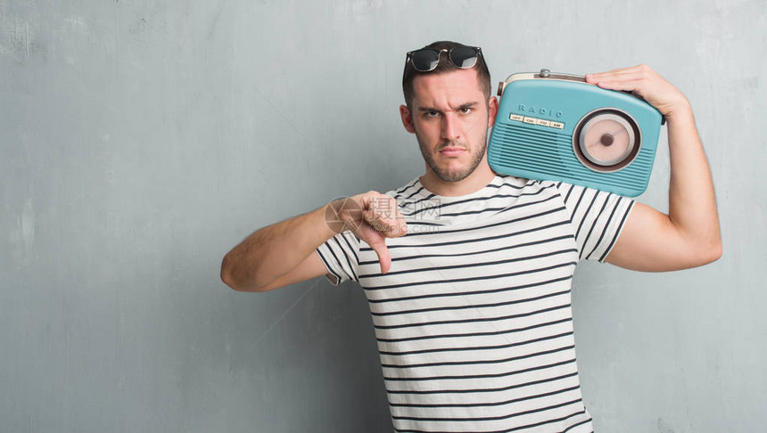 年轻的白人男子在灰色的格朗基墙上用愤怒的脸在老式收音机上听音乐图片