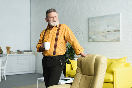 时髦的长胡子老人拿着一杯咖啡看远视而坐在家里手椅旁图片