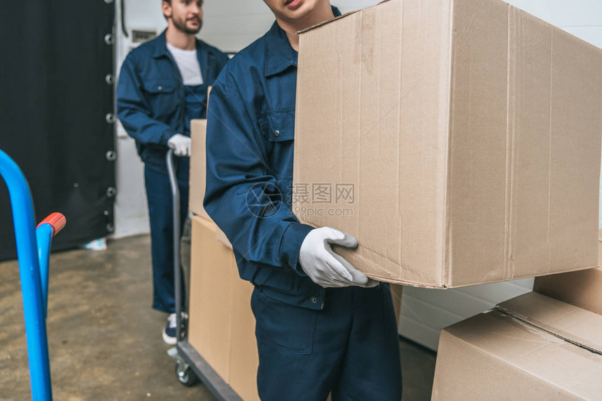 身穿制服携带纸板箱和仓库内手提卡车的两辆搬运车在仓库中图片