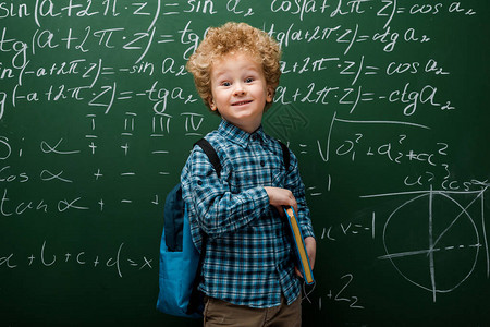 带着数学公式在黑板附近持有书的幸福儿童图片