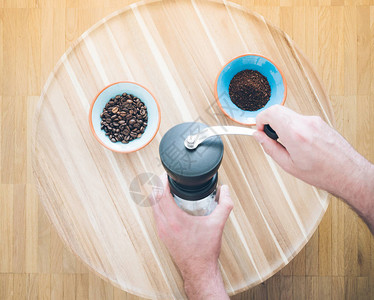 男手操作手动咖啡研磨机和两个装有咖啡豆的碗和木质表面研磨咖啡的男图片