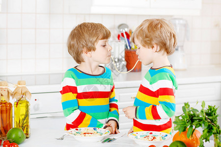 两个有趣的小男孩在室内厨房里吃长面条和新鲜蔬菜穿着五颜六色衬图片