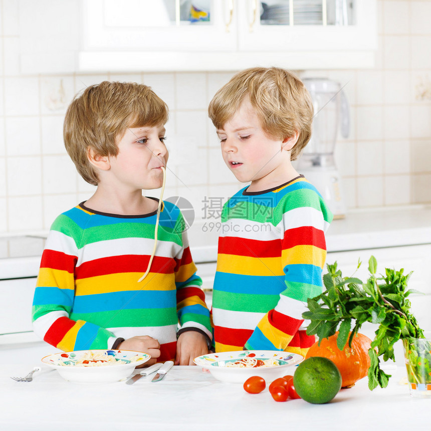 两个有趣的小男孩在室内的家用厨房里吃着面条和新鲜蔬菜图片