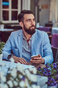 穿着时髦发型的胡子男近身肖像拿着智能手机坐在咖啡厅户外餐厅里图片