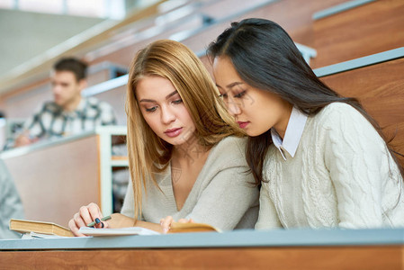 两名美丽的女孩在现代大学礼堂的书桌旁看笔图片