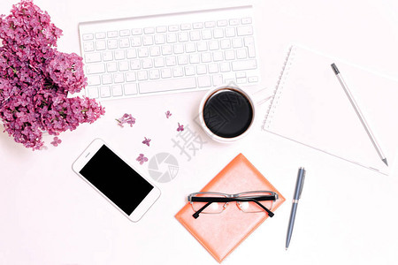 工作场所与电脑键盘手机眼镜咖啡杯笔记本铅笔钢笔和淡紫色花在白桌背景图片