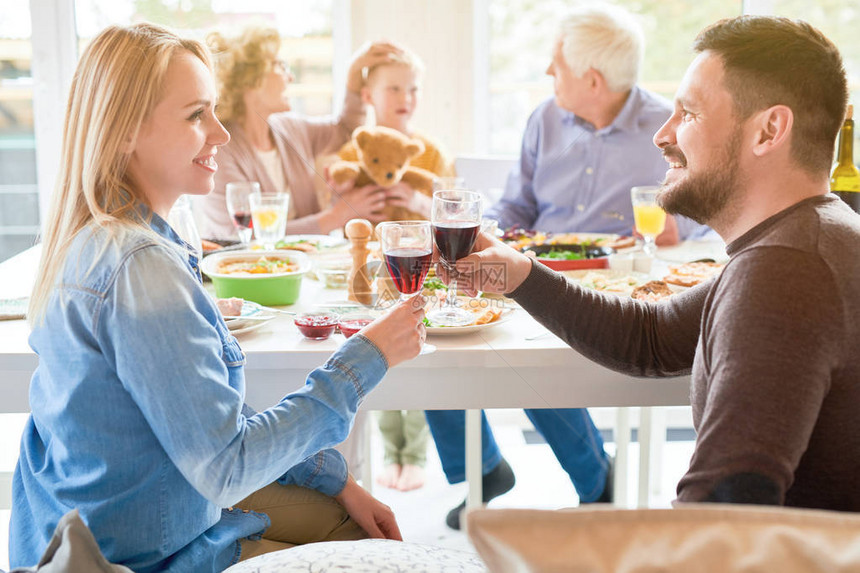 喜悦的现代夫妇在阳光下家庭晚餐时将葡萄酒杯连在一图片