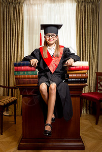 带着毕业帽子微笑的聪明女孩坐在书堆之间的桌边图片