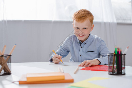 坐在桌边的红发学前男孩在课堂上绘画时带纸和彩色铅笔的快图片