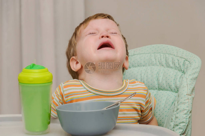 穿着条纹T恤的金发小男孩不想用灰色盘子里的勺子吃粥男孩在哭泣和抗拒图片
