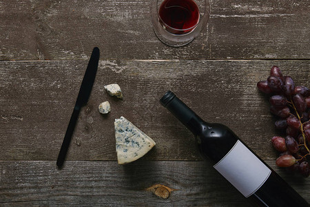 木桌上带空白标签的酒瓶带红酒奶酪葡萄和刀的玻璃图片