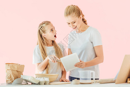 身穿白色T恤的幸福母亲和惊讶的女儿在用平板烹饪时使用粉红色孤图片