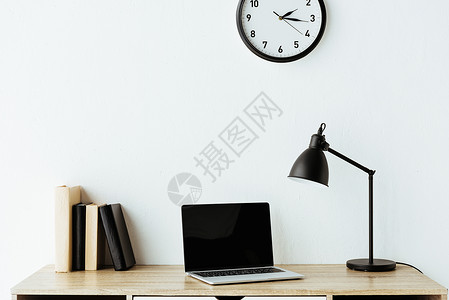 在白墙上挂着时钟的办公桌上放着书和台灯的笔记本电脑图片