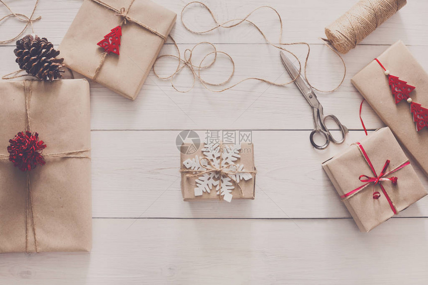 创意爱好用麻线丝带将圣诞假期手工制作的礼物包装在工艺纸中白色木桌上的剪刀和带弓和雪花的图片