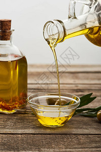从瓶中倒油到玻璃碗油瓶橄榄树叶和木背景图片