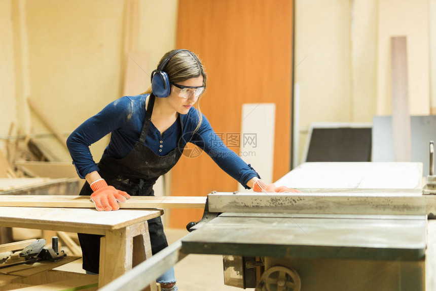 有吸引力的女木匠在使用木制车间的桌锯时佩戴耳图片
