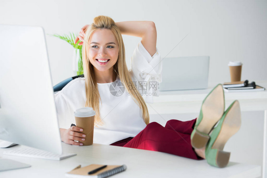 手持可支配咖啡杯和看办公室台式计算机的年轻女图片