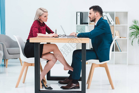 一对年轻的商务人士在办公室的桌子下一起工作和调情时互相看着图片