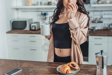 妇女在家中厨房的木制餐桌上与牛角面包和咖啡一起吃早图片