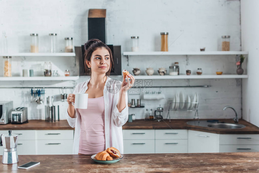 年轻妇女在家中厨房的木制桌边用牛角面包和咖啡与羊角面图片