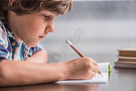 专注的小男孩坐在桌边时在书本上写图片