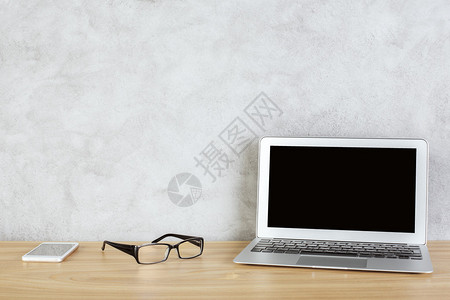 带纹理的混凝土墙背景的空白笔记本电脑屏幕眼镜和智能手机的木制桌面的图片
