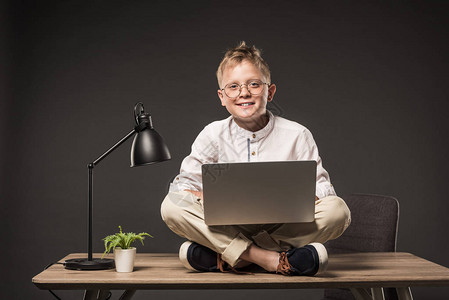 小男孩在用笔记本电脑坐在灰色底带植物和灯光的桌边时图片