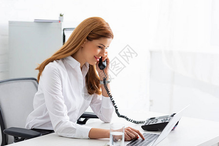 笑的女商人在电话上交谈和使用笔记本电脑在桌上说图片