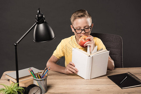 身着严重眼镜的男学生阅读书和在桌边用彩色铅笔钟表灯一叠书本和灰底背景图片
