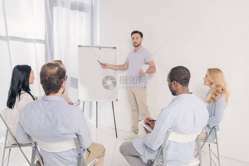 男心理治疗师指向白板和在治疗期间坐在椅子上的多种族图片