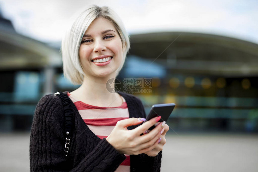 在火车站外使用手机时微笑的美丽年轻美女肖像图片