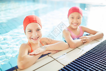 穿泳帽和游泳服的跨文化女孩在游泳池看相机时快乐地享受背景图片