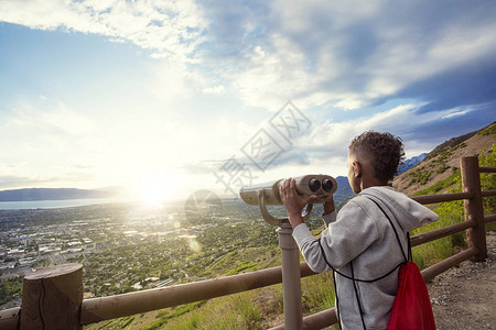 小男孩在徒步登山后透过望远镜向下俯视着城市的山区景图片