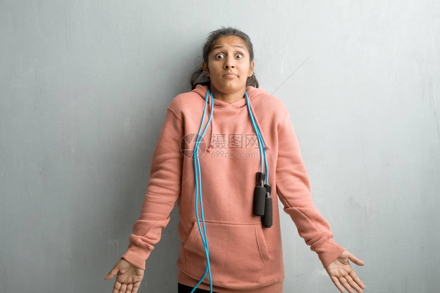 年轻的运动型印度女人靠墙怀疑和耸肩图片